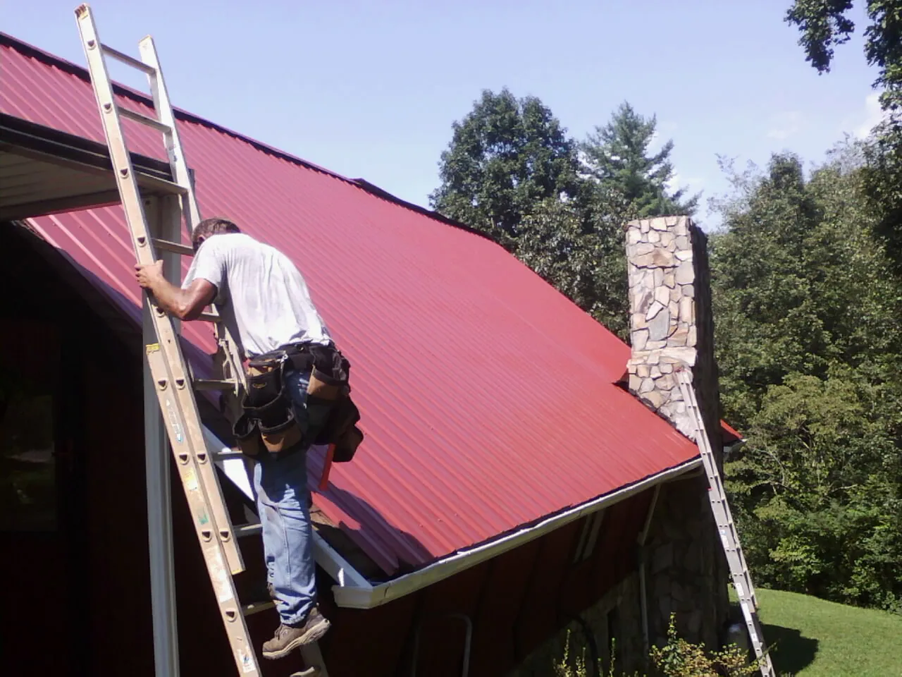 Roofer on a ladder beside a red metal roof and stone chimney — Soffit & Fascia Repair by Roofing Brothers in Colliersville